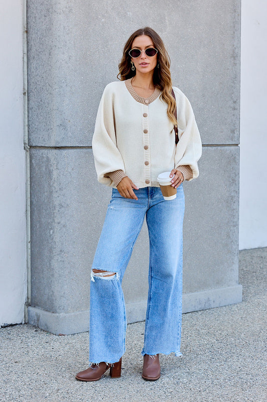 Woman in cream Contrast Ribbed Cardigan, sunglasses, ripped jeans, and brown boots holds coffee by a gray concrete wall.