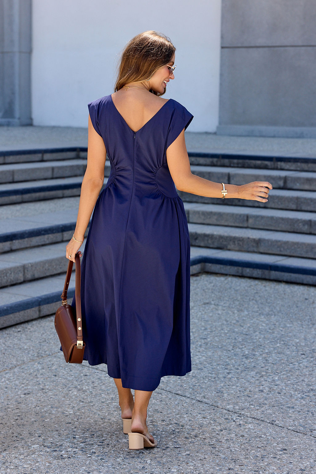 Woman in a Navy Ruched Detail Midi Dress with beige sandals holds a brown handbag and wears sunglasses on concrete steps.