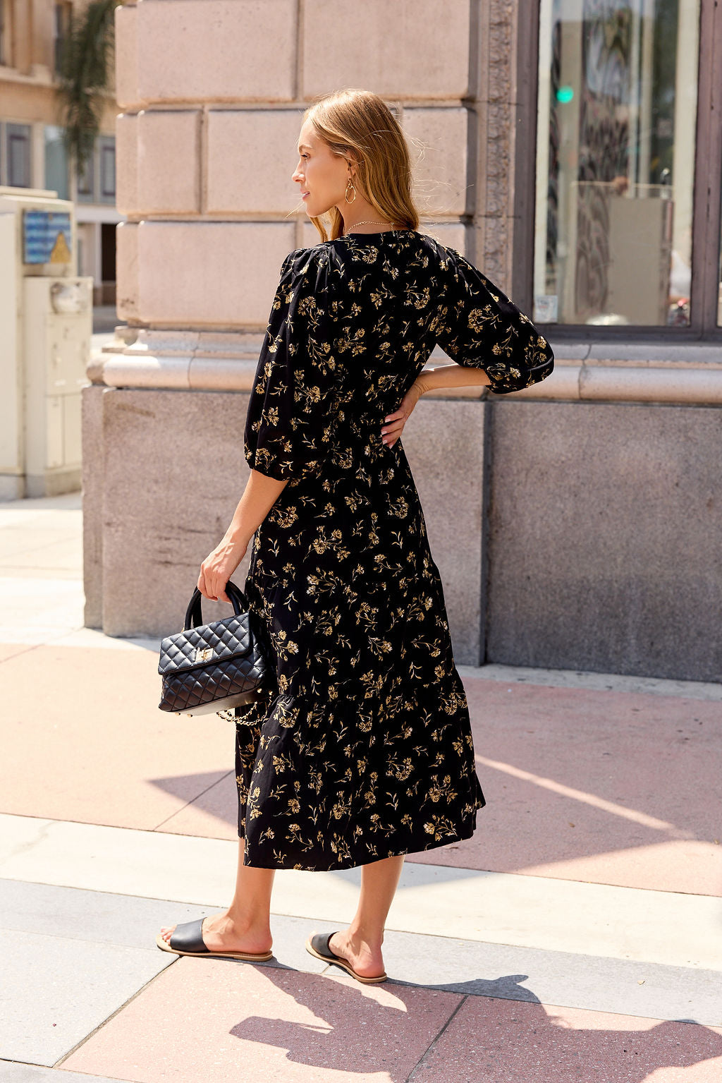 Woman in Ember Floral Midi Dress - Black with sandals holds a black quilted handbag on a sunny sidewalk.