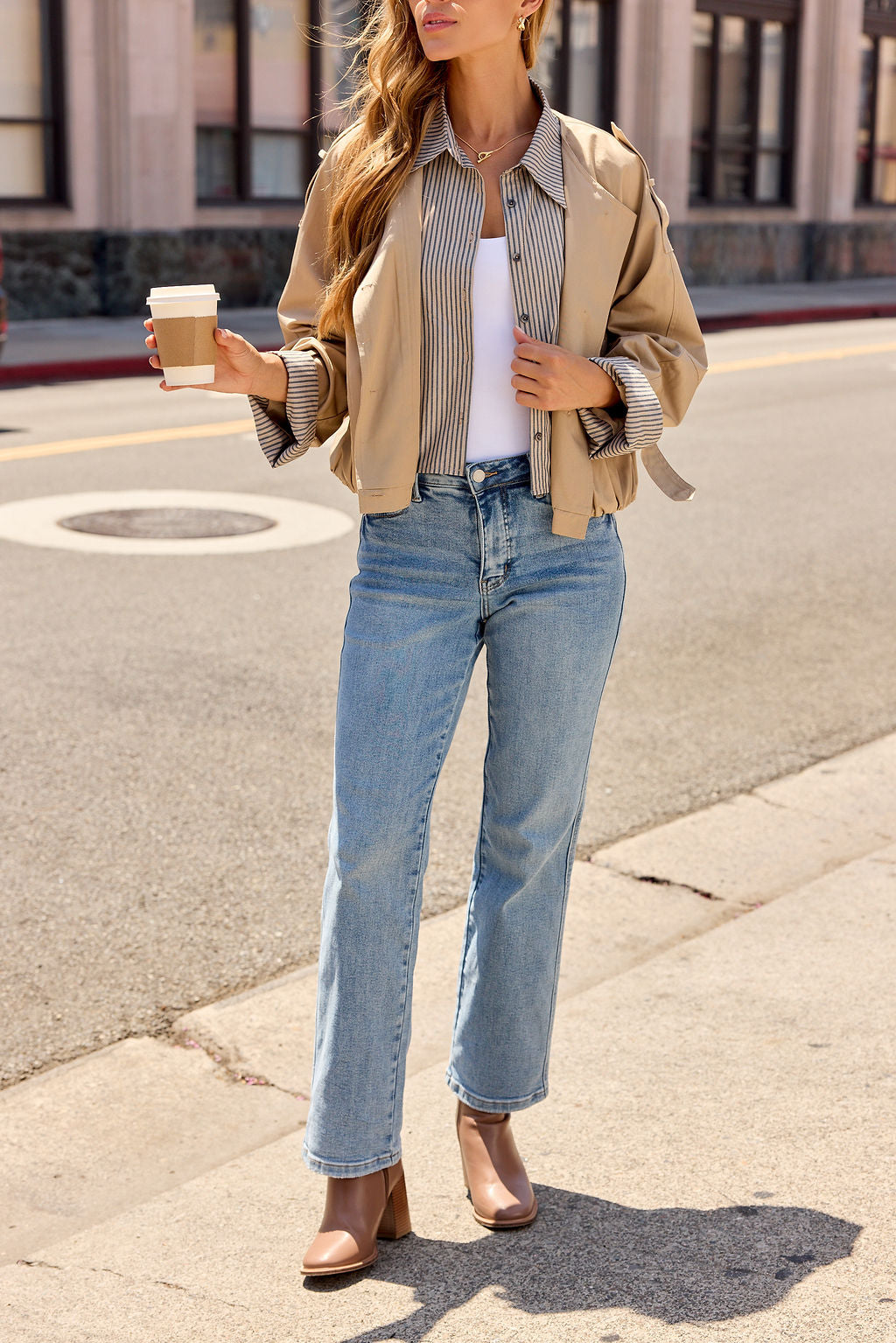 Woman in a Contrast Striped Shirt Jacket - Camel, white top, jeans, and boots holding coffee on a city sidewalk.