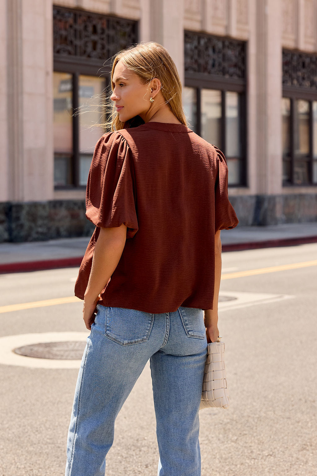 Woman wears a Puff Sleeve Tie Front Top in brown with blue jeans, holding a white handbag, back to the camera on the street.