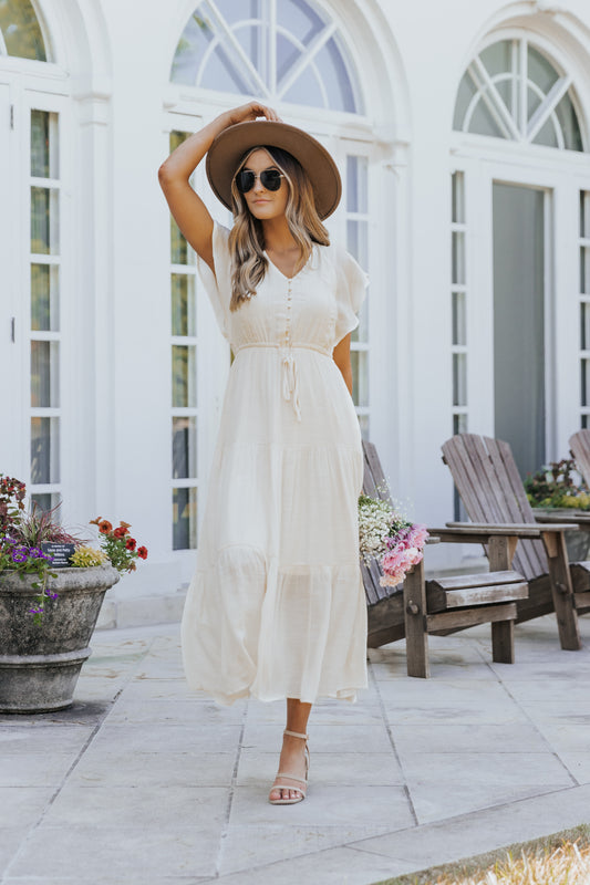 Woman in a Celine Crochet Lace Tiered Boho Midi Dress and hat walks on a patio with wooden chairs, flowers, and arched windows.