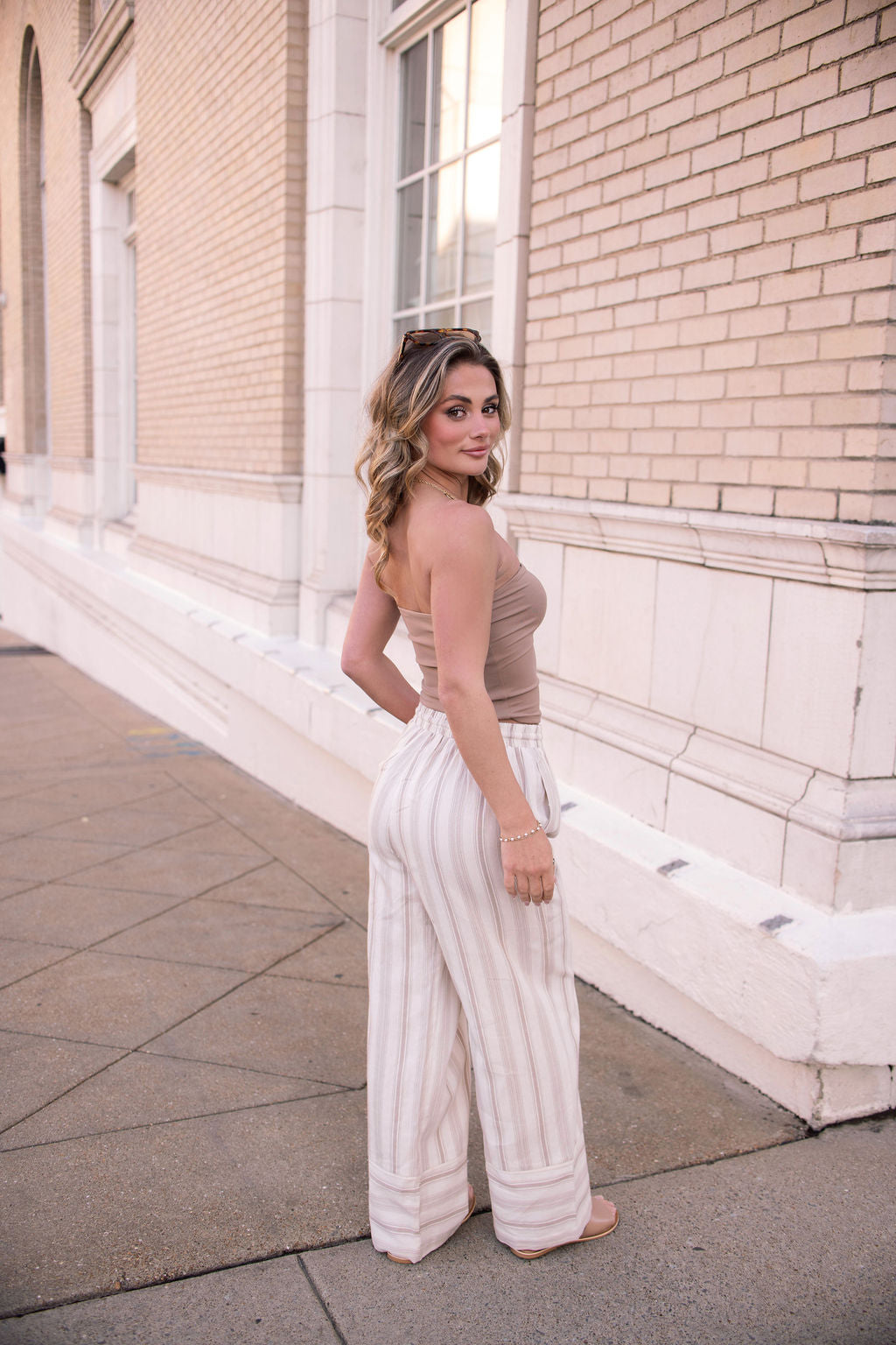 Woman on a sidewalk wears Mocha Striped Palazzo Pants - FINAL SALE, showing off their wide-leg silhouette as she looks back.