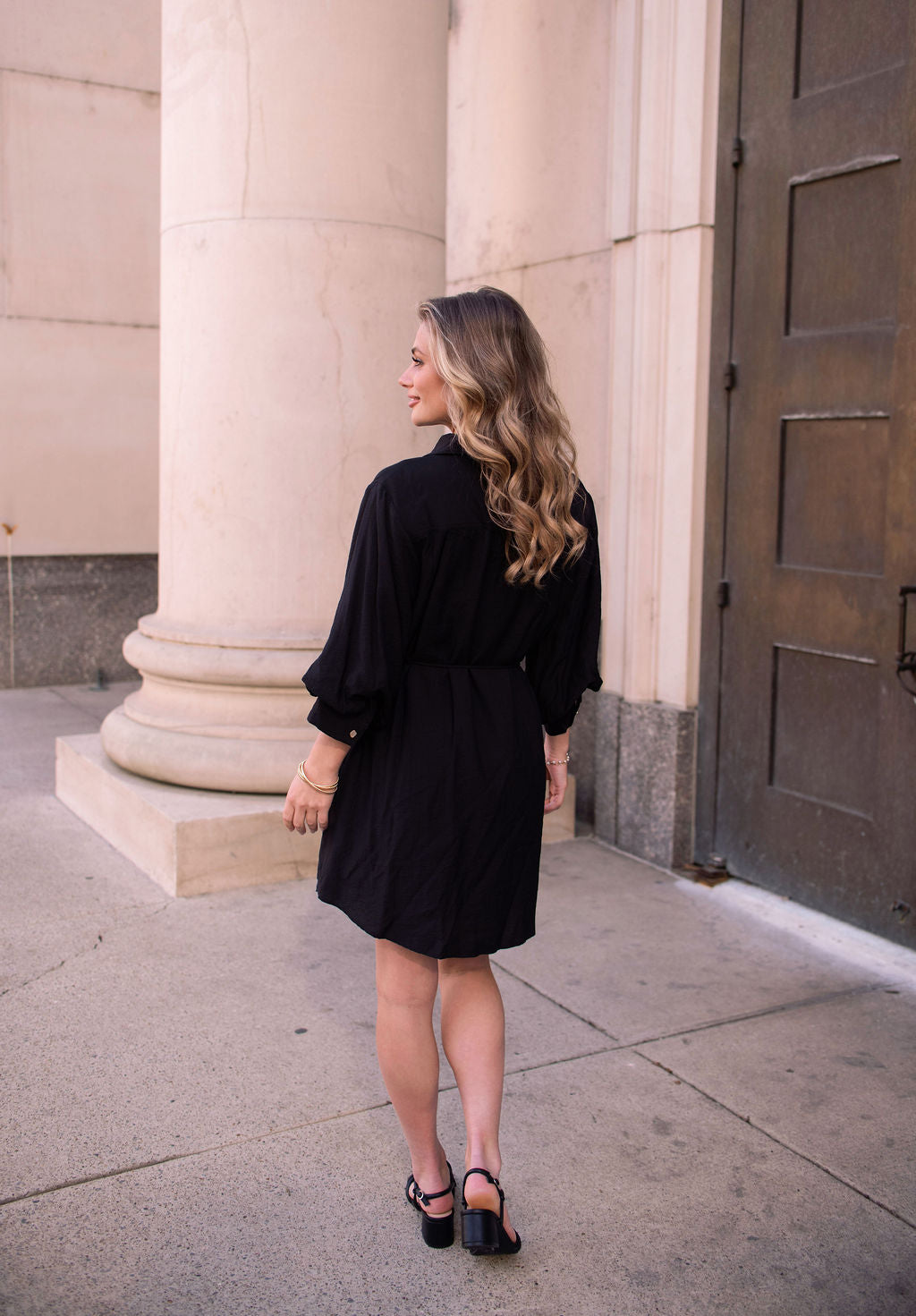 A woman in the Black Tie Mini Shirt Dress and sandals stands outdoors by large columns, facing away from the camera.