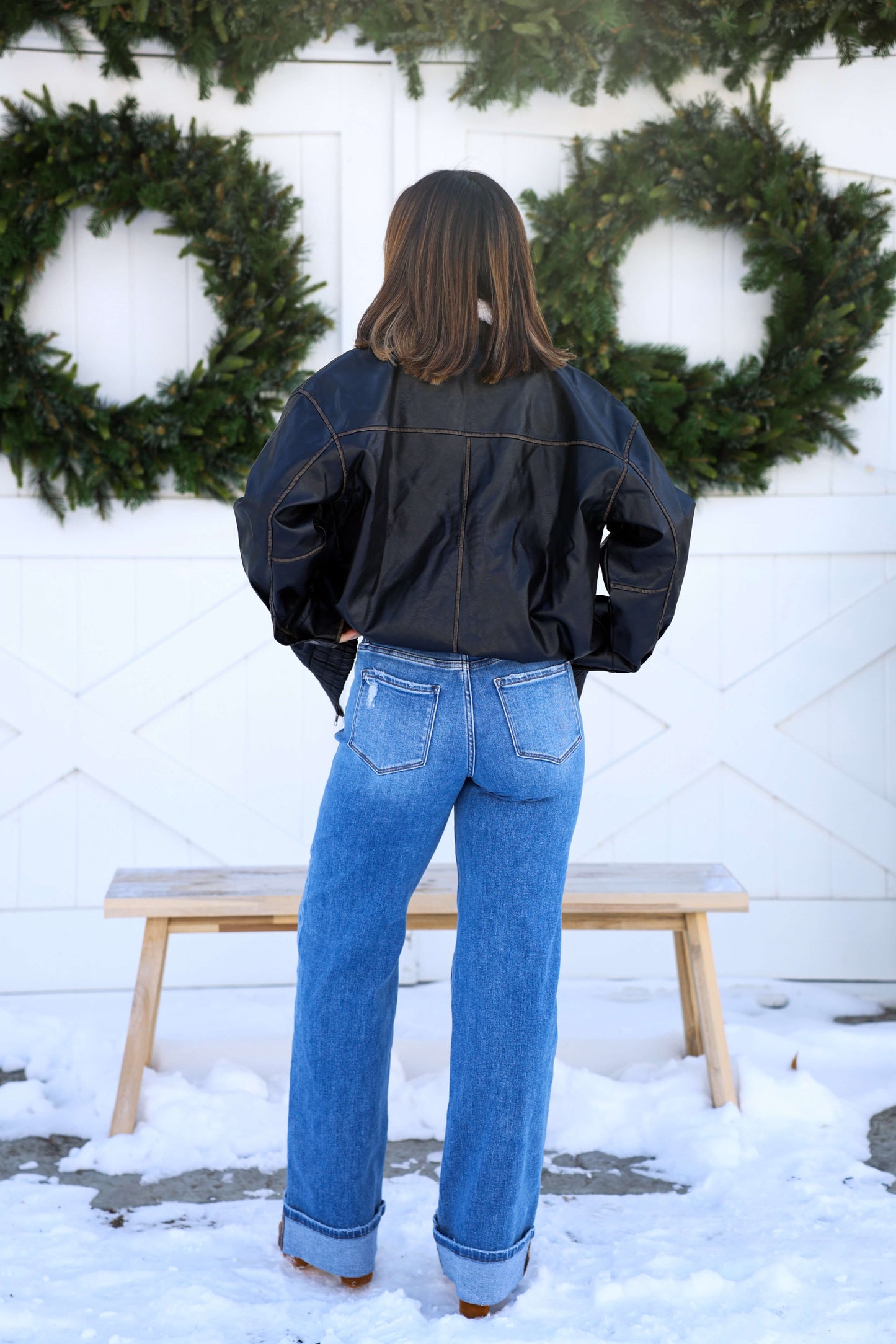 Someone wearing Vervet Medium Wash High Rise Cuffed Jeans stands on snow, facing away, with green wreaths on a white wall.
