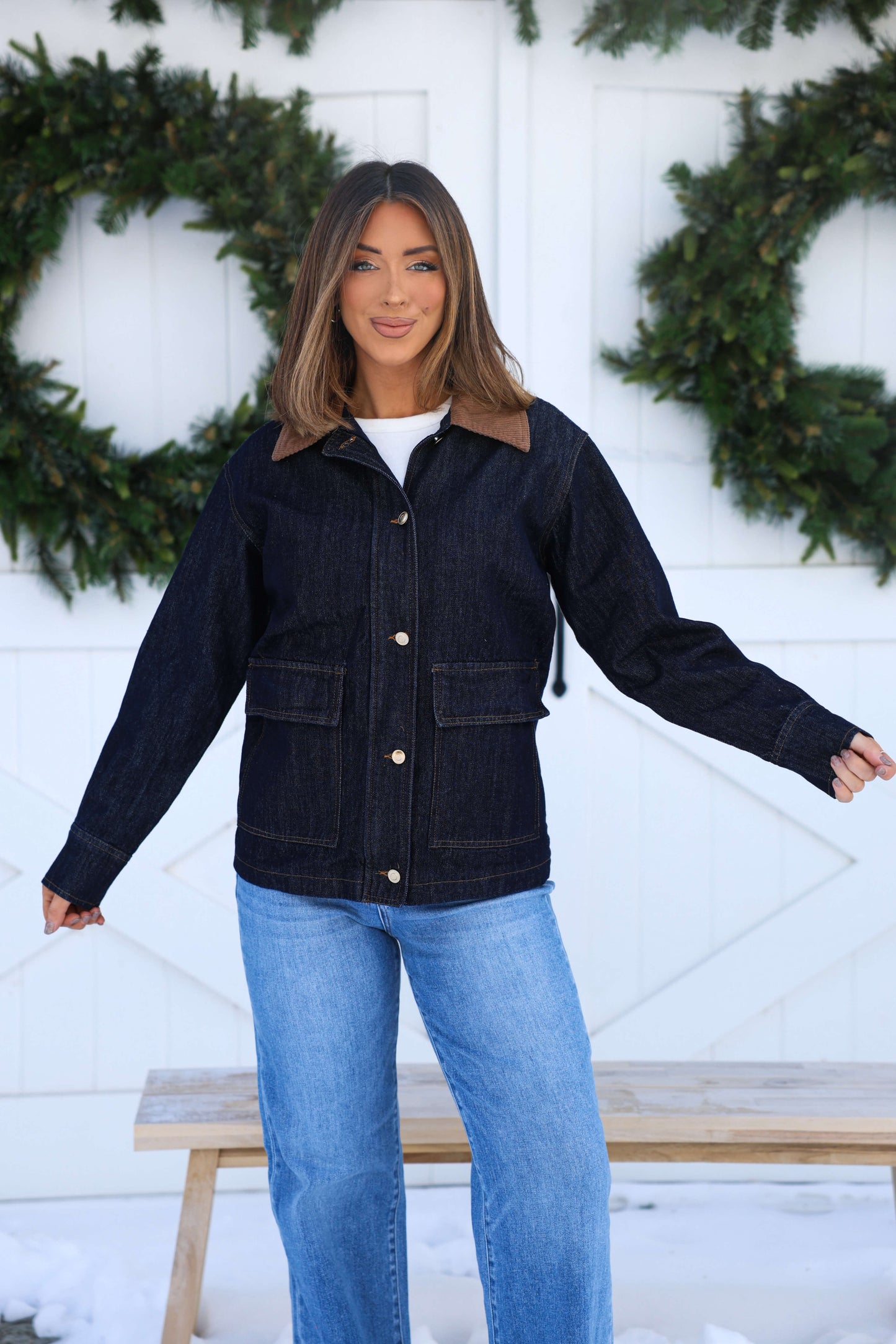 Woman smiles in front of white wall with wreaths, wearing a Dark Denim Carpenter Jacket and matching jeans.