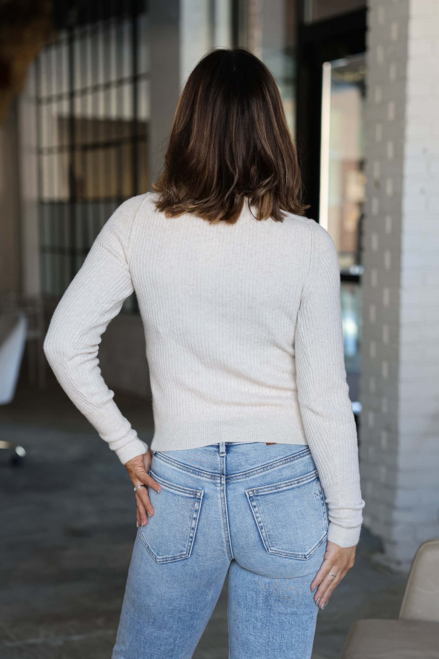 A woman stands indoors, facing away, wearing the Free People Oatmeal Ever After Pullover and light blue jeans.