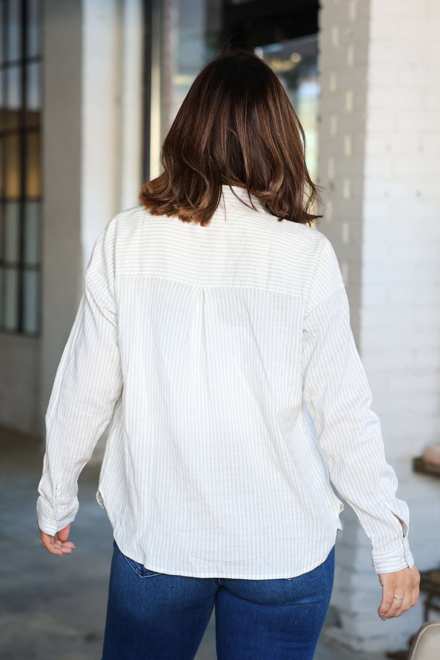 A person with shoulder-length brown hair wears the Thread and Supply Edison Tan Striped Shirt and blue jeans, facing away indoors.