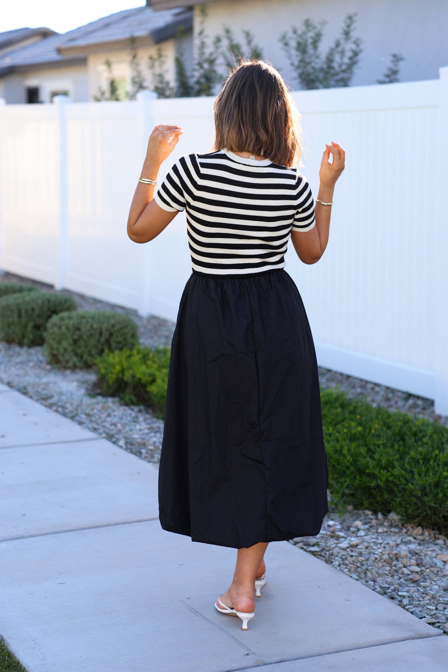 Woman wearing a Black Stripe Bubble Hem Midi Dress walks on a sidewalk by a white fence and greenery, facing away from the camera.
