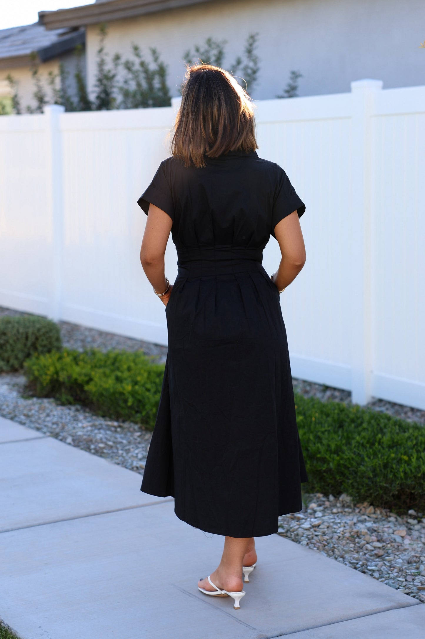 A woman in a Black Double Belted Midi Dress and white heels stands on a sidewalk near a white fence and greenery.