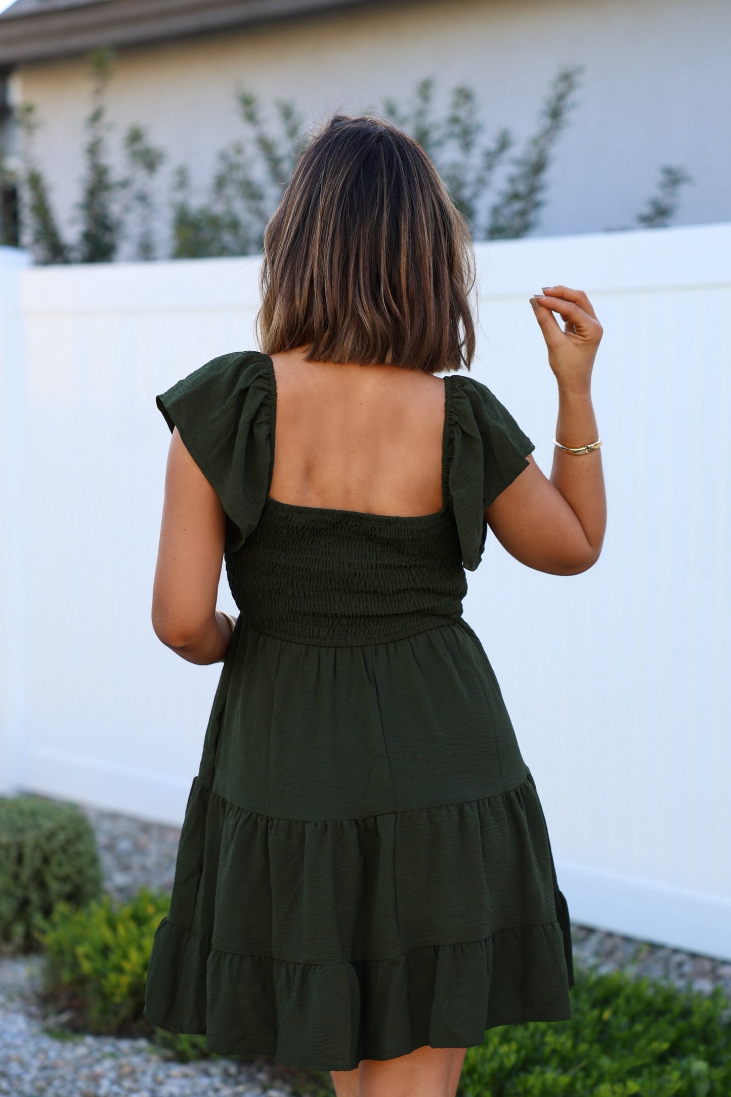 A woman in Ava's Sweetheart Olive Mini Dress stands outdoors, facing a white fence and lush garden.