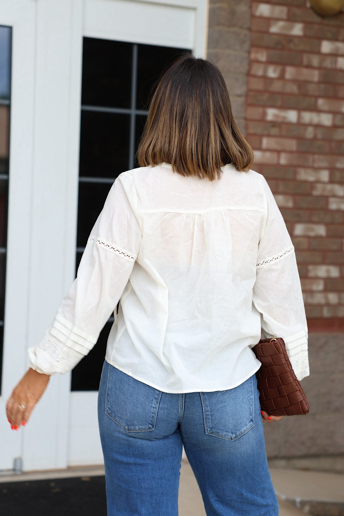 A woman seen from behind wearing an Off White Crochet Long Sleeve blouse and blue jeans, holding a brown woven clutch.
