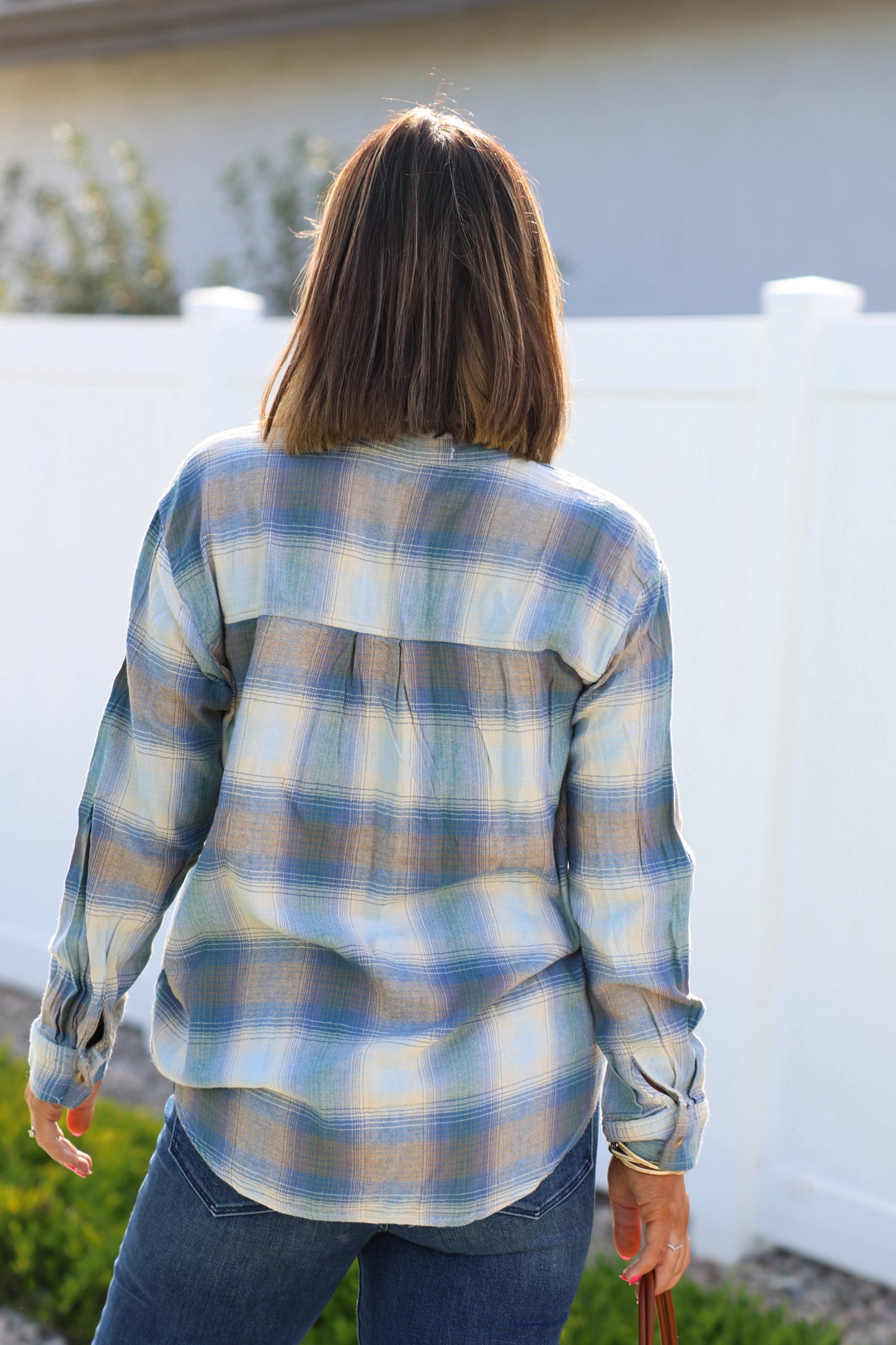 A person with shoulder-length hair in a Blue Plaid Button Down Flannel Shirt and jeans stands outside facing a white fence.