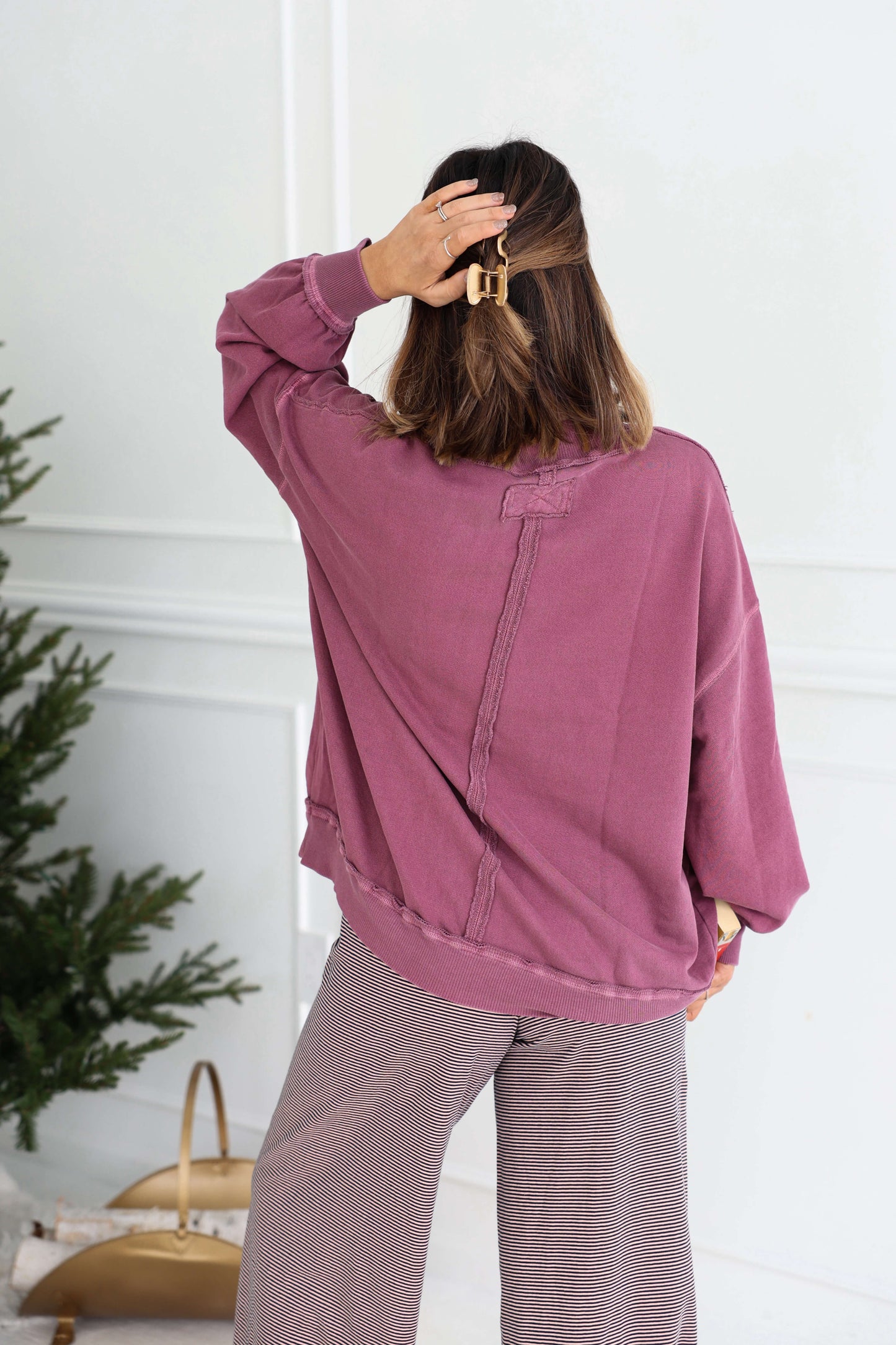 A woman in a Free People Iris Orchid Palm Springs Pullover and gray pants stands with her back to the camera, hand in her hair.