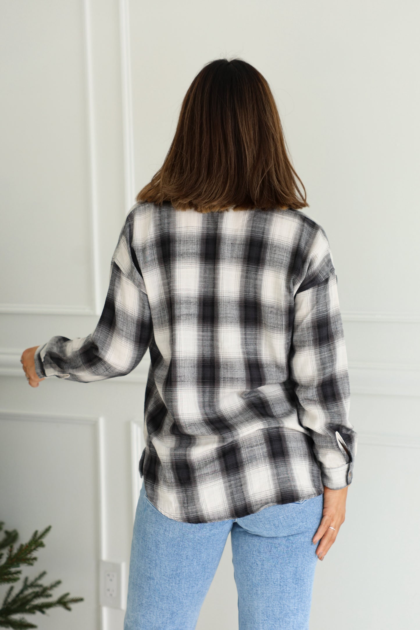 A woman with shoulder-length brown hair models the Madelyn Charcoal Plaid Flannel Shirt and light blue jeans, back to the camera.