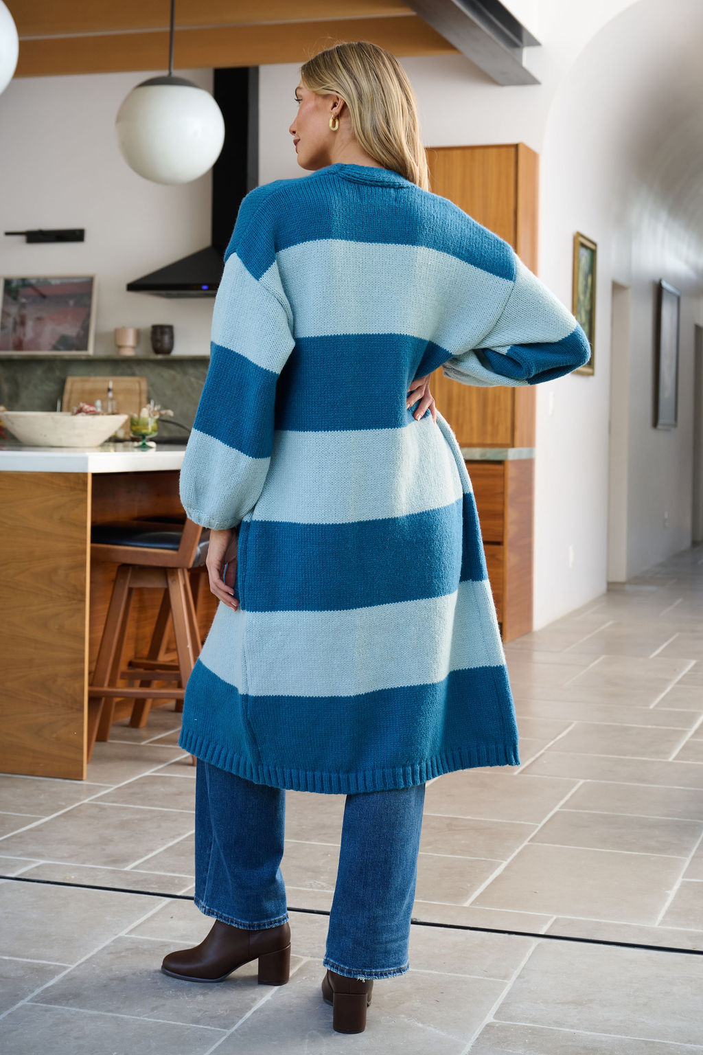 A woman stands in a kitchen wearing a Blue Striped Open Front Duster Cardigan, blue jeans, and brown heeled boots—winter ready.