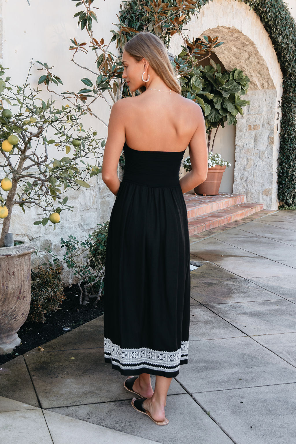 Woman in a Black Strapless Embroidered Midi Dress stands outside by a potted lemon tree, facing away from the camera.