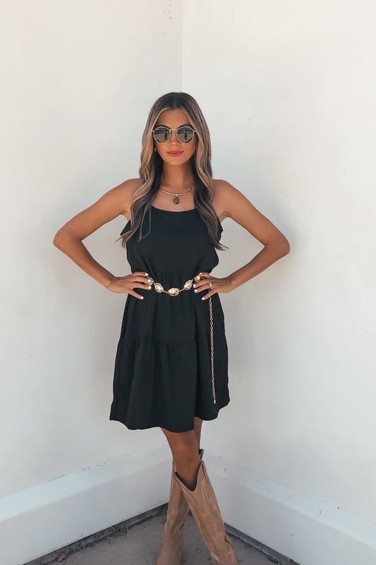 Woman in On Point Tiered Mini Dress - Black with adjustable straps and comfort stretch, sunglasses, and tan boots against a white wall.