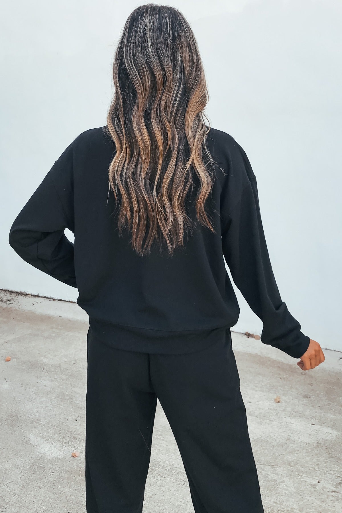 A person with long, wavy hair stands outside, wearing the Ultra Soft Brushed Fleece Sweatshirt in black and matching pants.