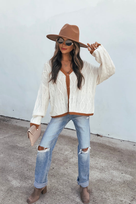 Woman in hat, sunglasses, Cream Contrast Trim Cardigan, ripped jeans, and boots poses outdoors holding a clutch against a plain background.