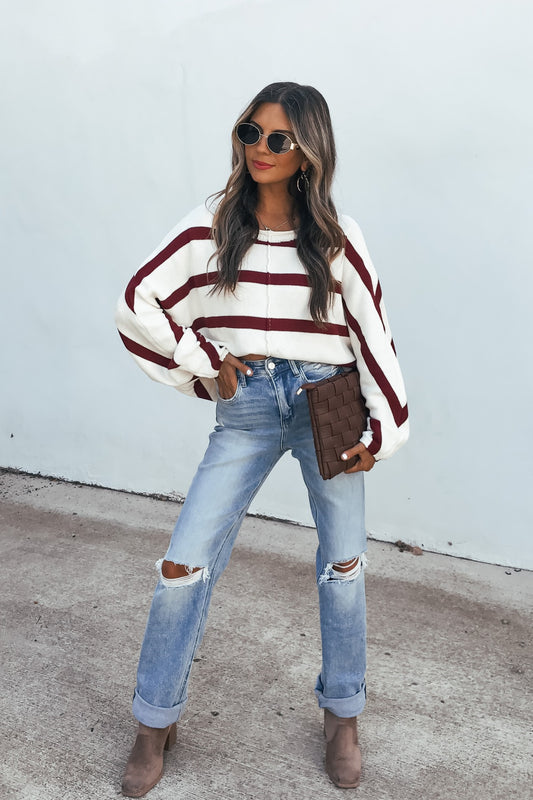Woman in a Burgundy Striped Dolman Sleeve Sweater, ripped jeans, and brown boots stands against a white wall.
