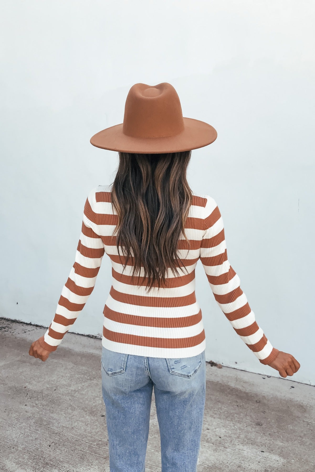 Someone in an Ivory Striped Long Sleeve Top and blue jeans stands facing a light wall, arms out, wearing a brown hat.