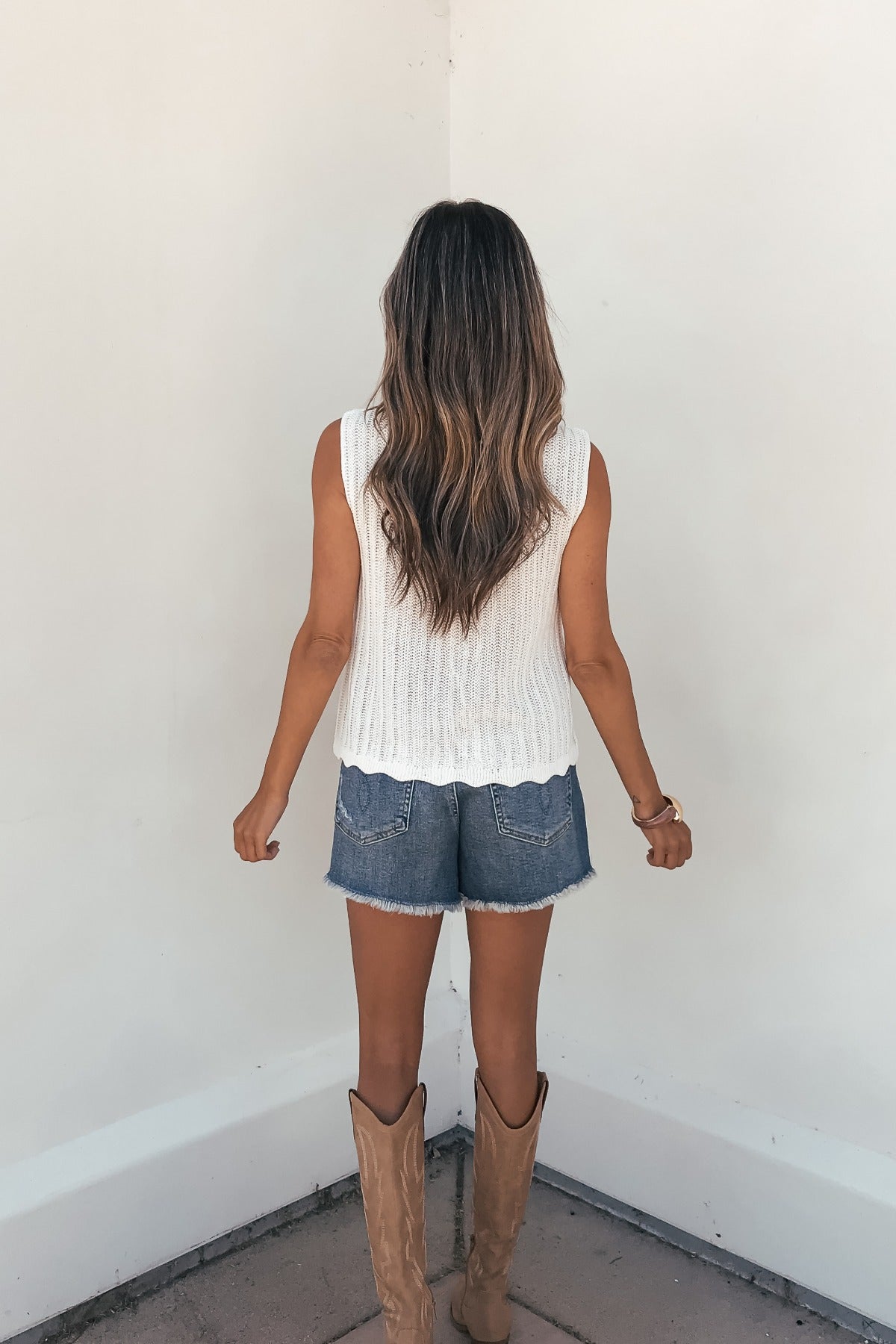 Woman with long wavy hair wears the Sunlace Tie Front Sweater Vest in cream, facing a white wall.