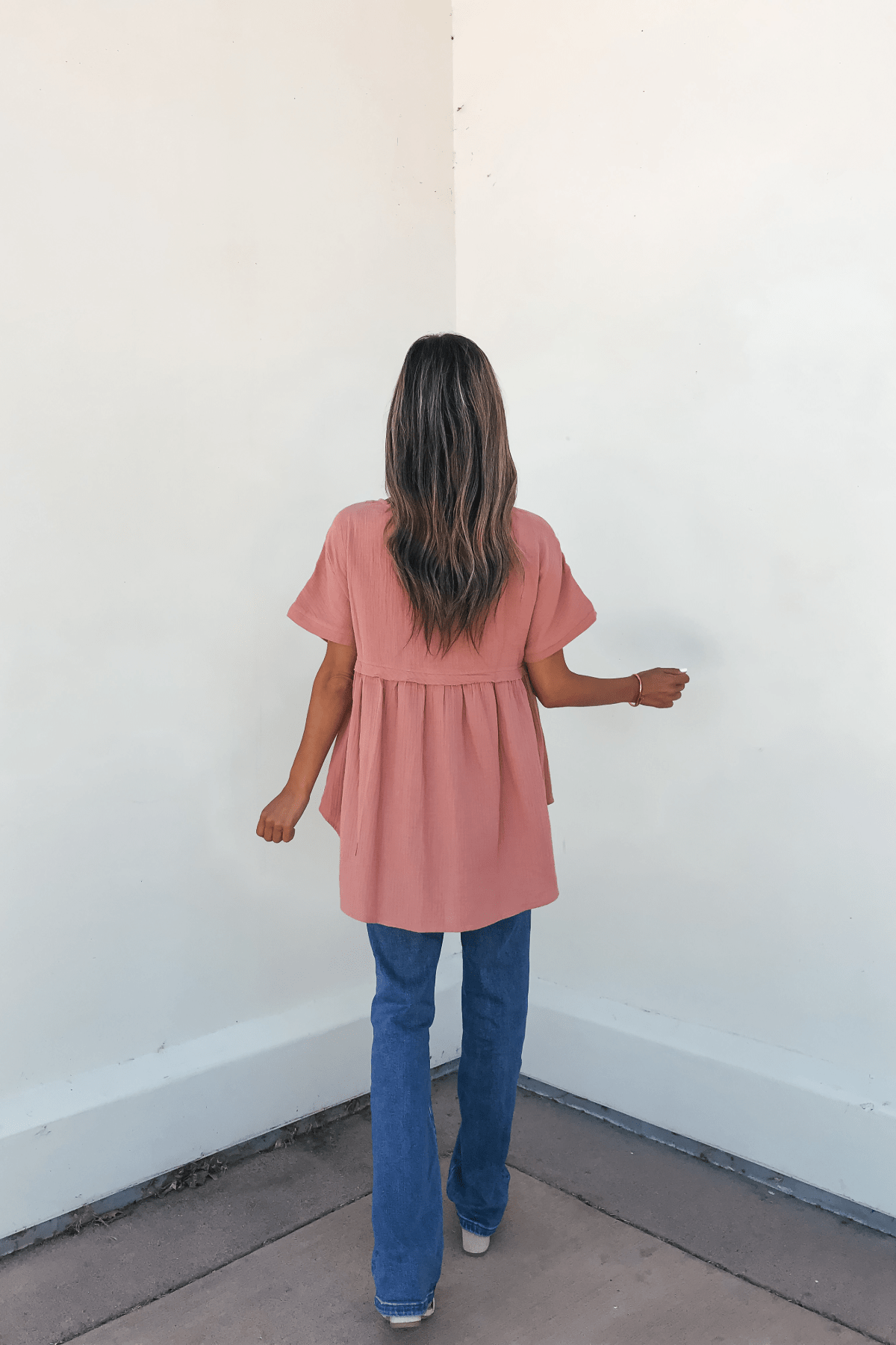 A woman in the Addison Coral Pleated Tunic Top and blue jeans stands with her back to the camera, highlighting this wardrobe essential.