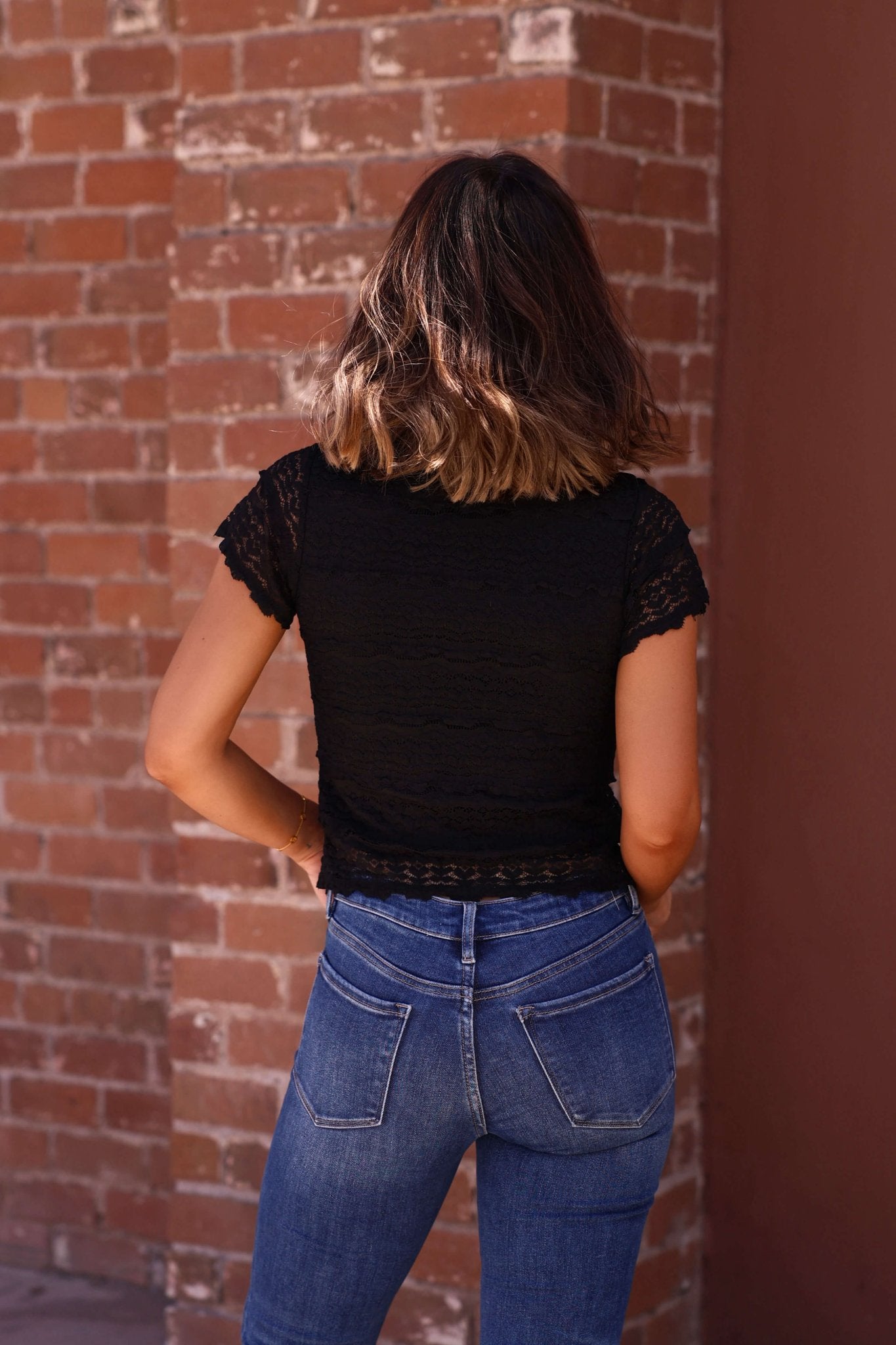 A woman in the Amara Black High Neck Lace Top - DOORBUSTER and blue jeans stands with her back to a brick wall.