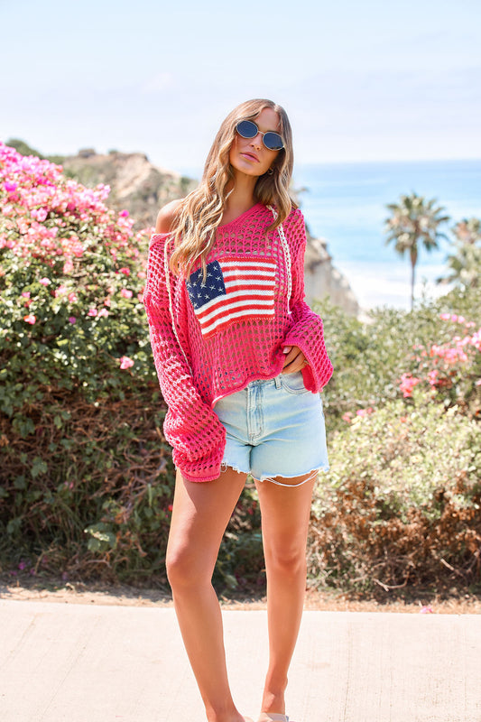 Woman wearing the All American Open Knit Hoodie and denim shorts by beachside flowers, enjoying the sun.