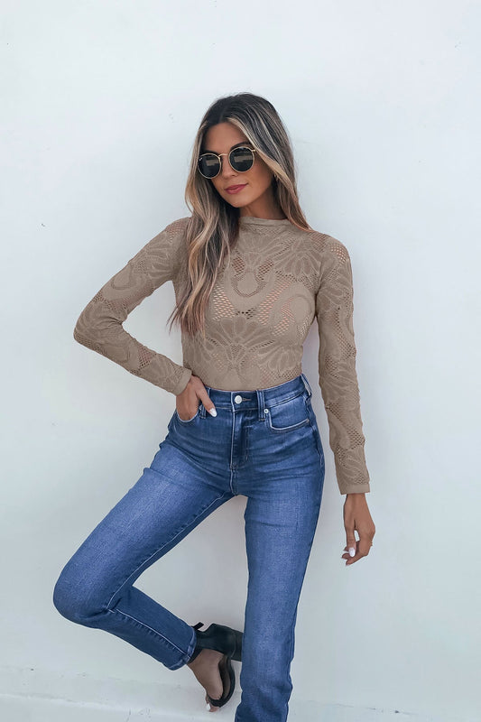 Woman wearing sunglasses, Ash Mauve Mock Neck Fishnet Top, and high-waisted jeans poses against a white wall with one hand in pocket.