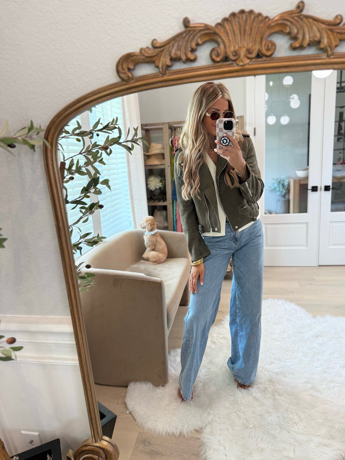 Woman in sunglasses takes a mirror selfie wearing the Audrey Olive Green Vegan Suede Moto Jacket; her dog sits on a beige chair.