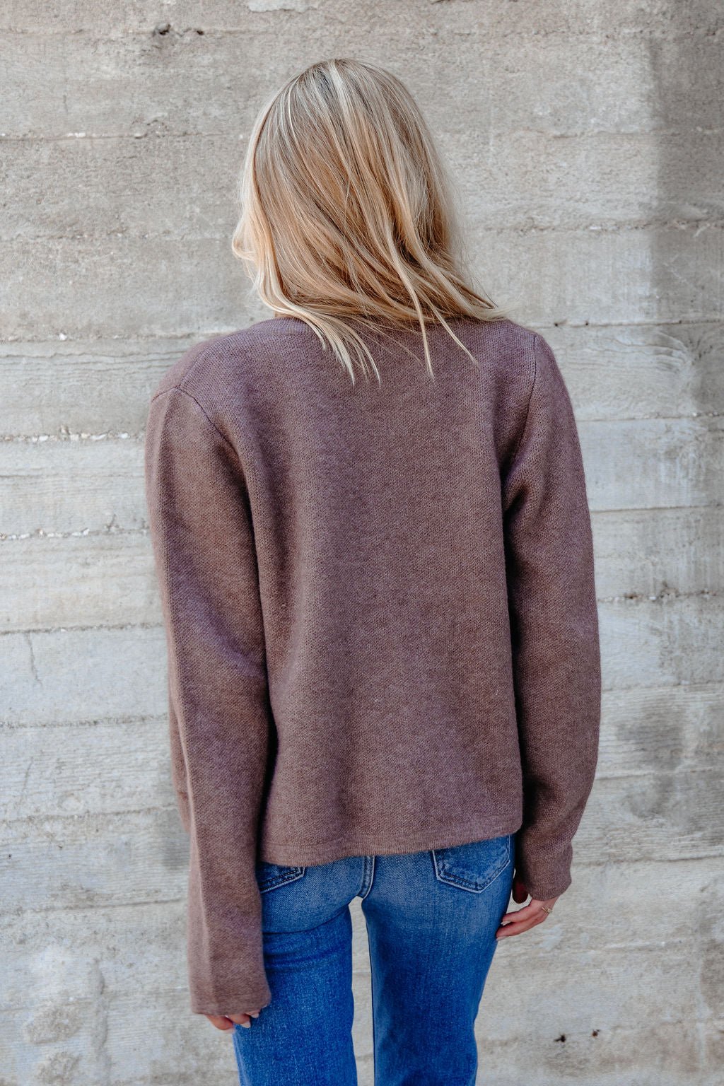 A person with blonde hair wears the Autumn Button Front Sweater Cardigan - Brown and blue jeans, facing a concrete wall.