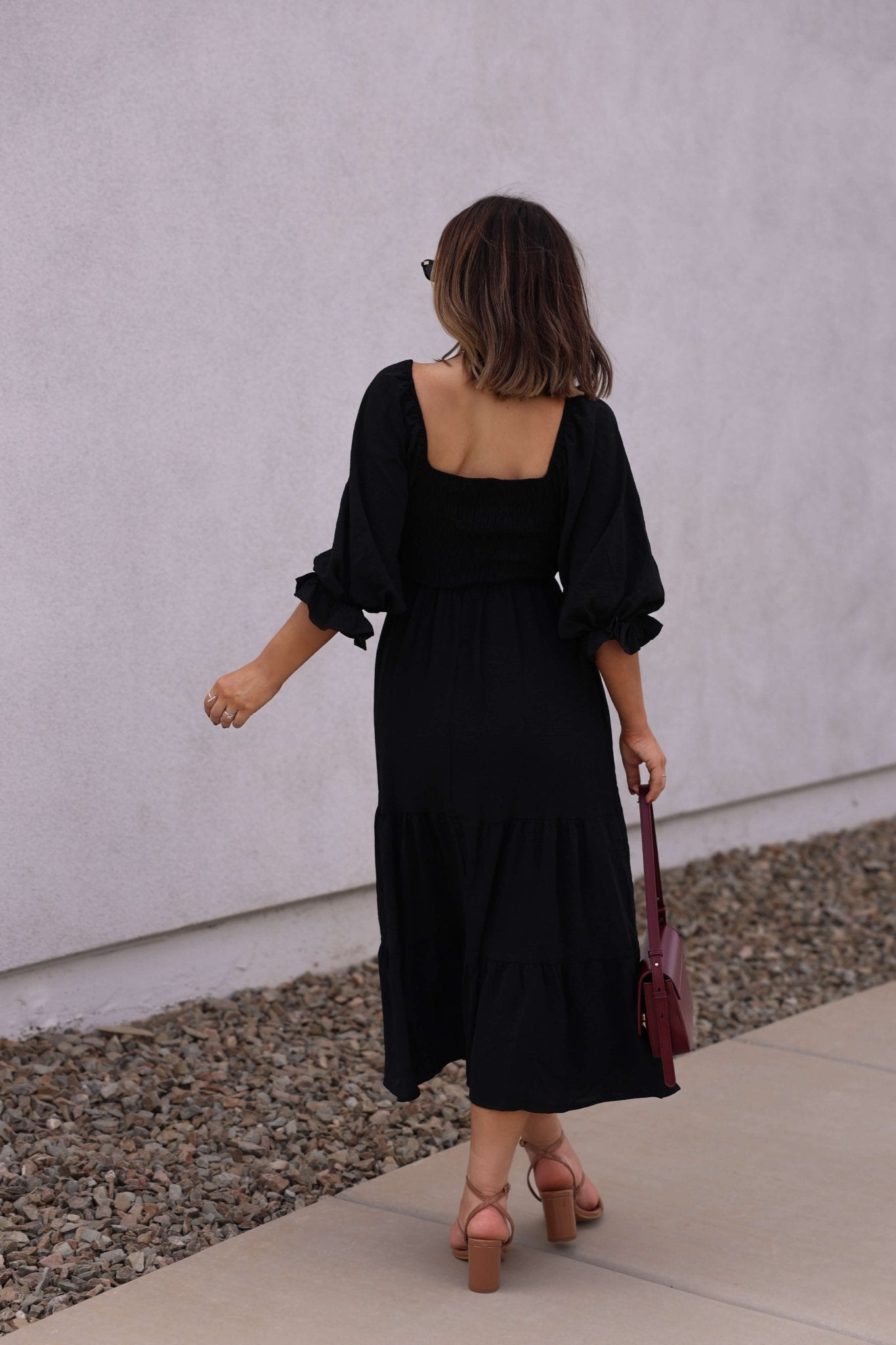 A woman walks on a sidewalk, seen from behind, wearing the Autumn Nights Black Tiered Midi Dress and holding a maroon handbag.