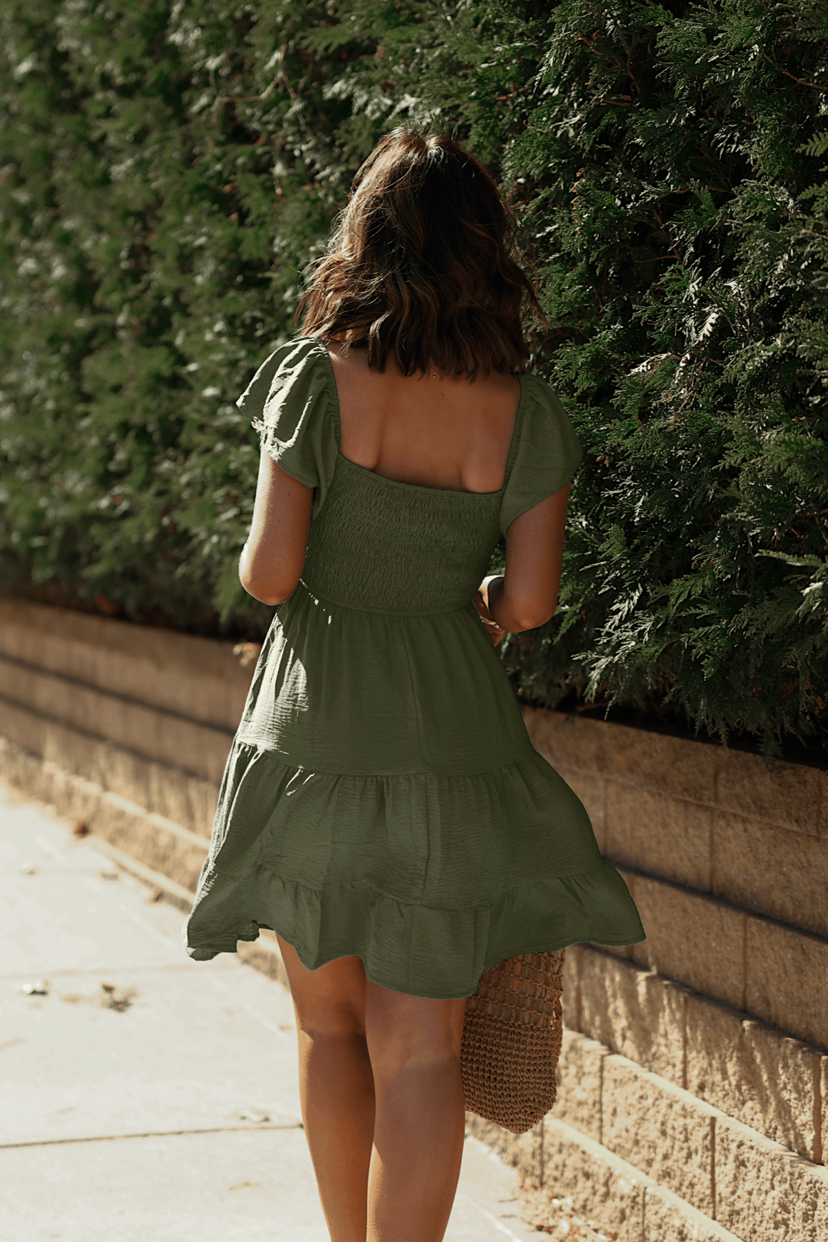 A woman in Ava's Sweetheart Olive Mini Dress walks along a sunlit sidewalk next to a green hedge and brick wall.
