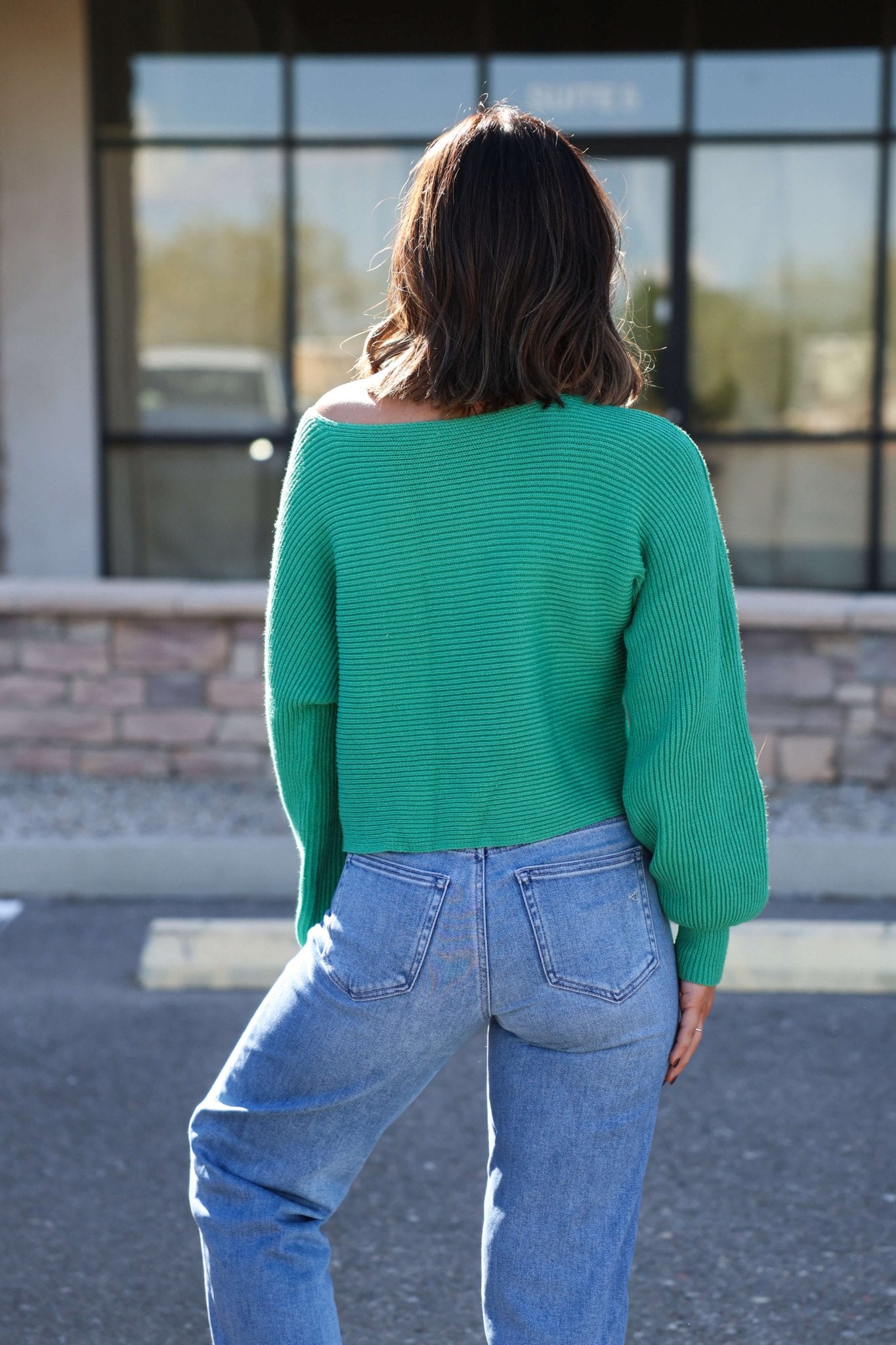 Woman with wavy brown hair wears the Avery Ribbed Boat Neck Sweater in green, paired with blue jeans, facing away outdoors.