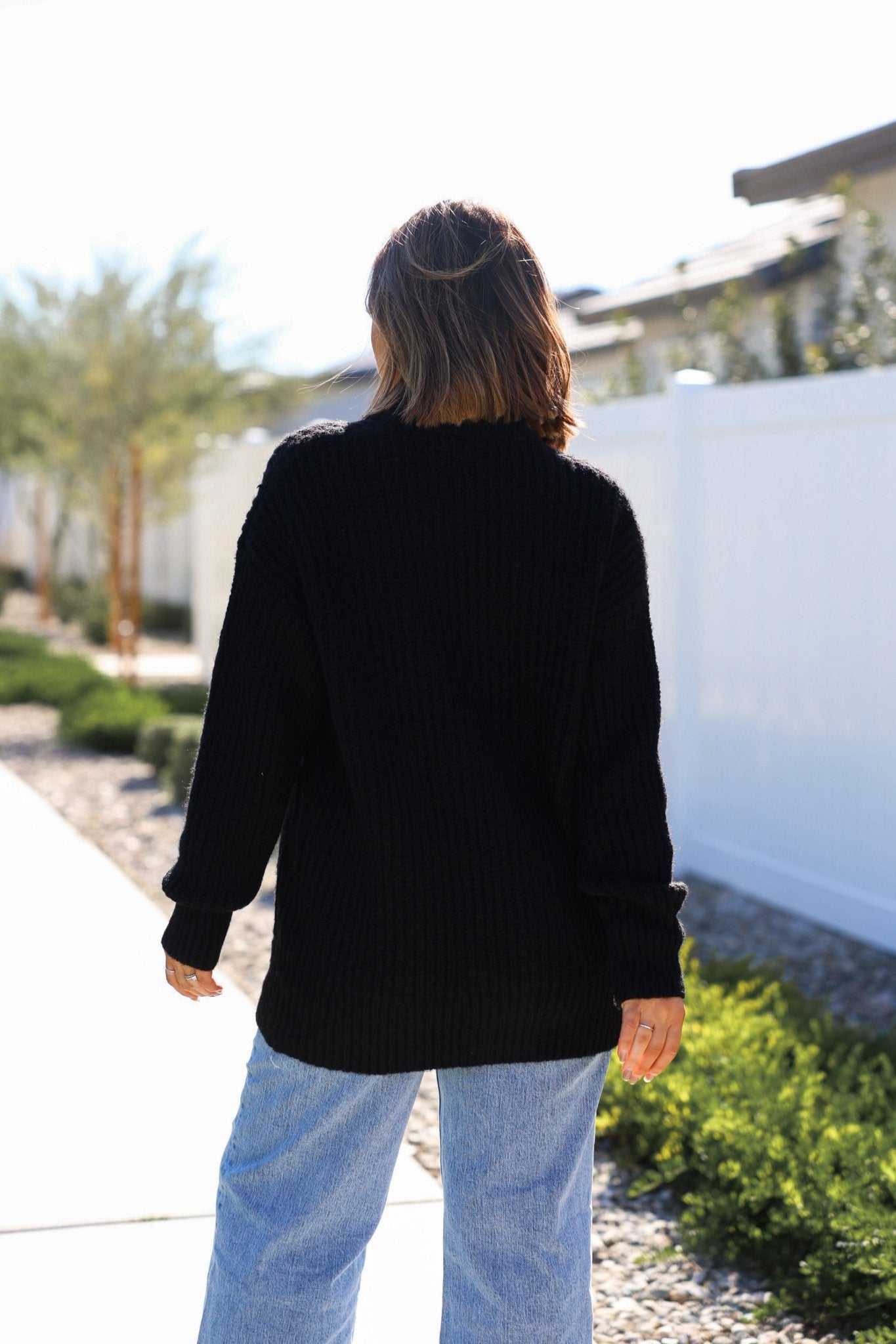 A person with shoulder-length hair wears the Black Heavy Knit Button Down Cardigan - FINAL SALE, standing outside on a sunny day.