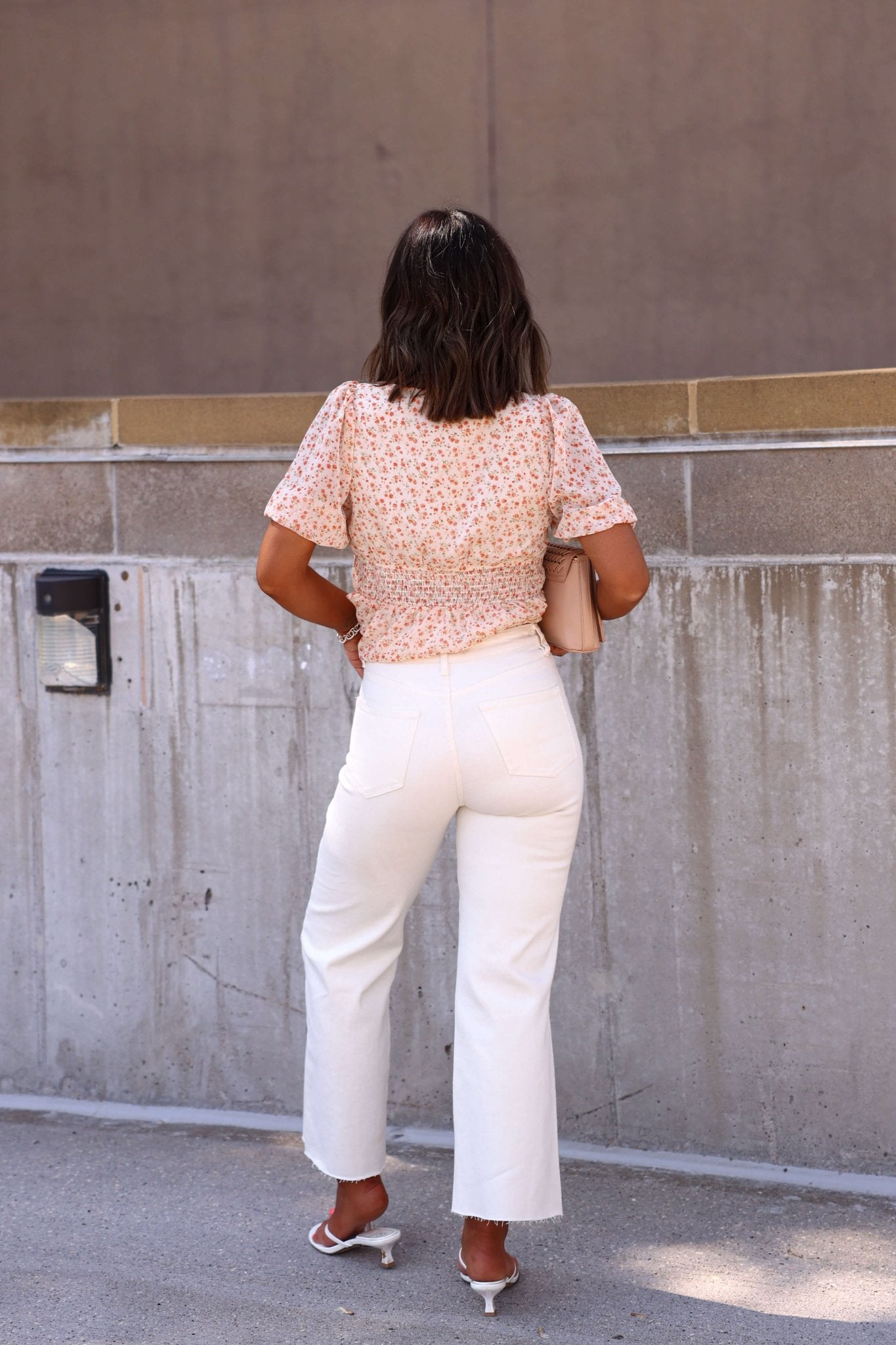 A woman in a floral blouse and Bayeas Cream High Rise Wide Leg Jeans holds a beige clutch, facing a concrete wall.