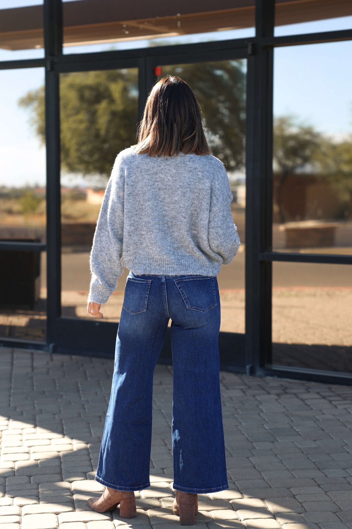 A woman in a gray sweater and Bayeas Lisa Dark Wash Wide Leg Jeans stands outside on a stone path, facing away.