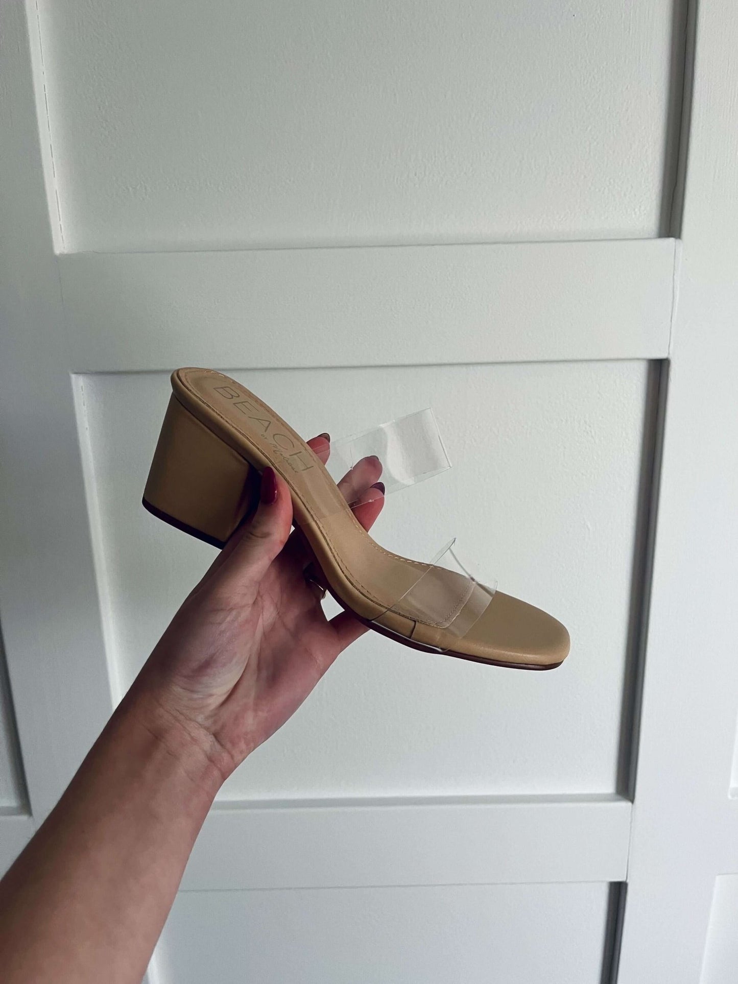 A hand holds the Beach by Matisse Aura Nude Strappy Heels with clear straps in front of a white paneled wall.