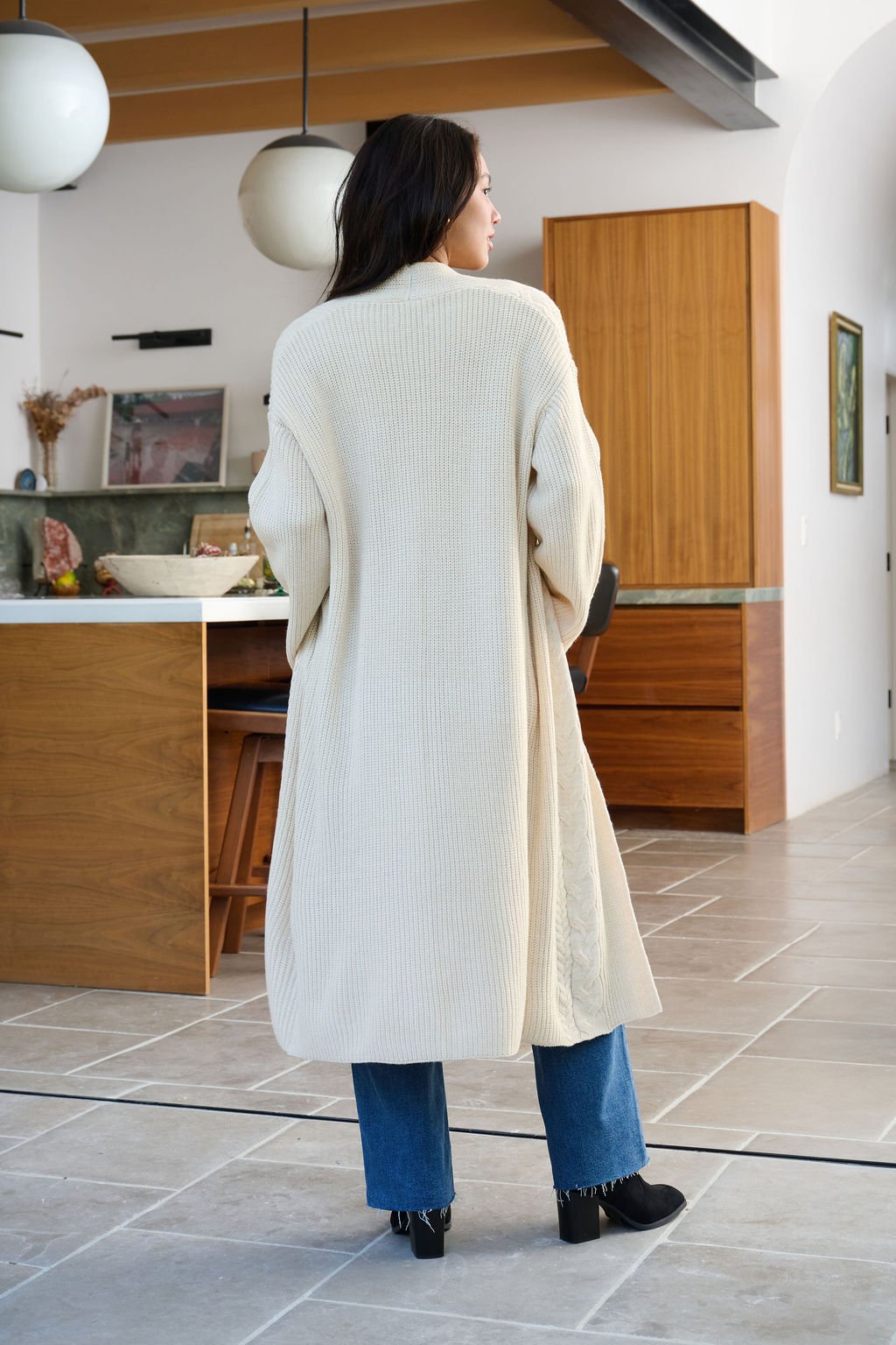 A woman wears a Beige Ribbed Open Front Duster Cardigan with blue jeans and black shoes, standing in a modern kitchen, facing away.