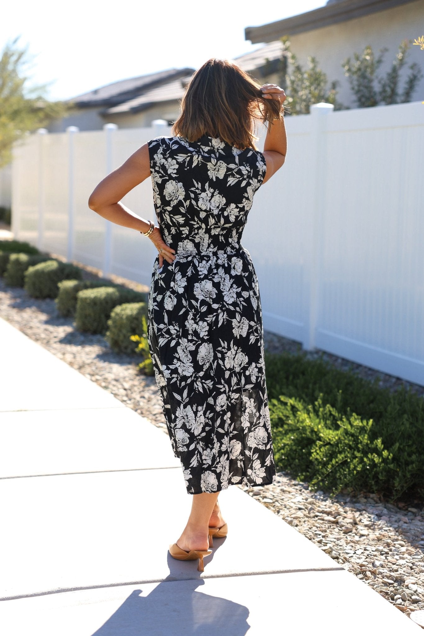 A woman models the Black and White Floral Print Smocked Midi Dress outdoors, with a white fence and greenery behind her.