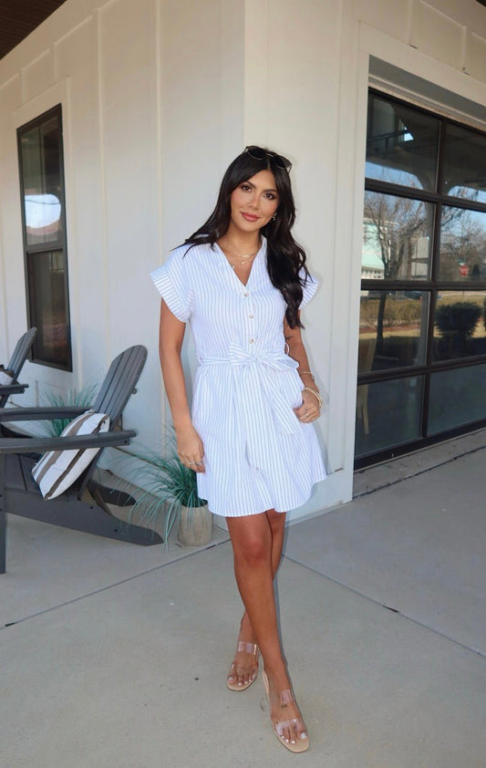 Woman standing outside a modern building in a Black and White Pinstripe Mini Dress and sandals, with long dark hair.
