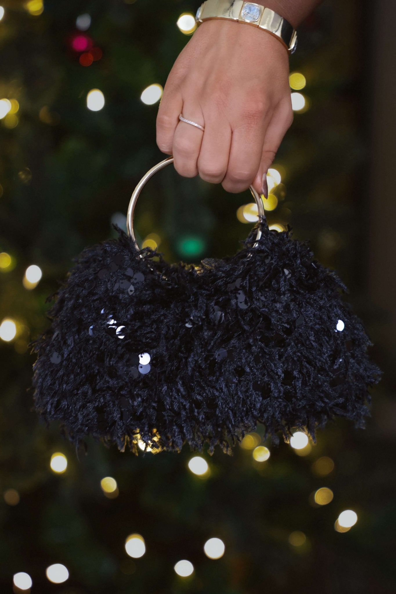 A hand holds the Black Charlotte Sequin Evening Bag with a gold handle before blurred festive lights.