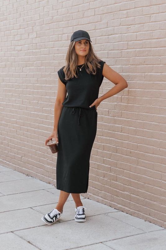 Woman in a Black Drawstring Slit Midi Dress, cap, and sneakers holds an iced coffee by a light brick wall.