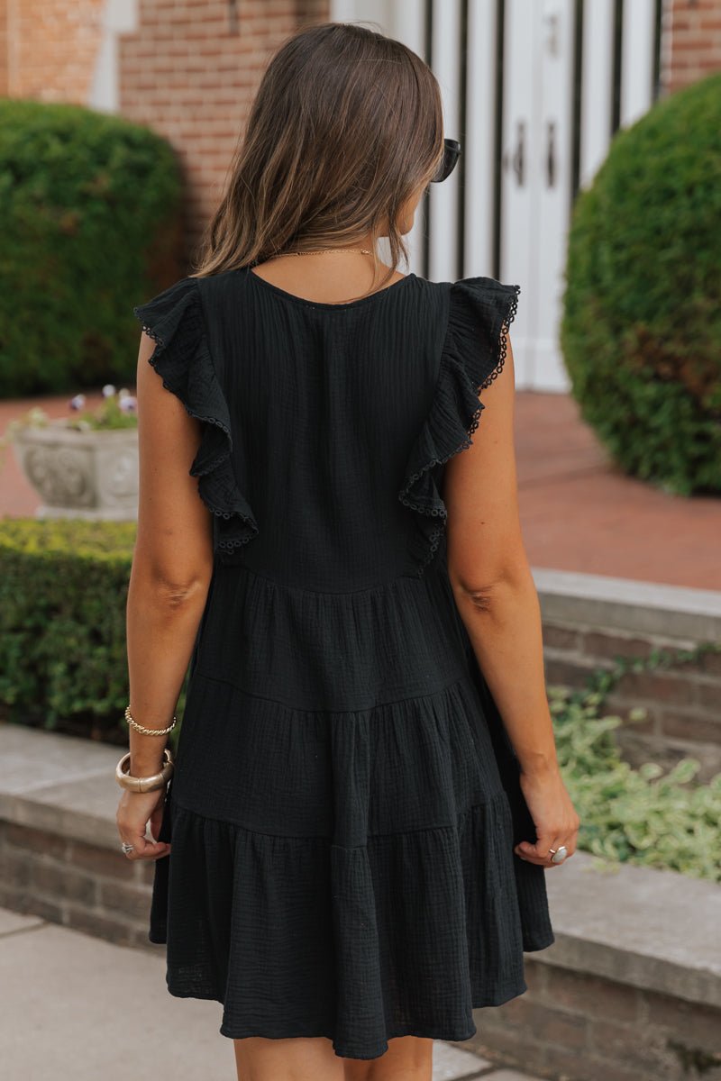 A woman in a Black Embroidered Tiered Mini Dress - DOORBUSTER stands outside with greenery and buildings behind her.