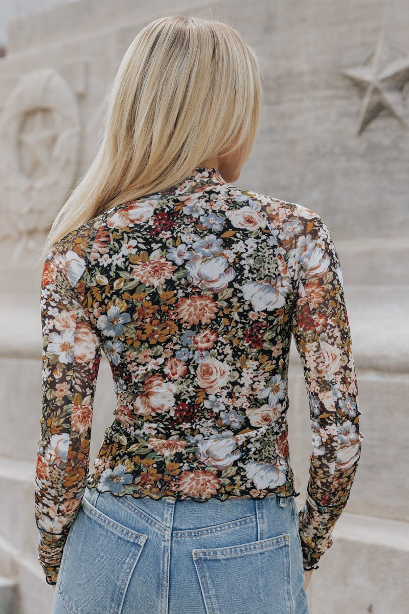 A woman with long blonde hair wears the Black Floral Mesh Top - DOORBUSTER and blue jeans, outdoors with her back to the camera.