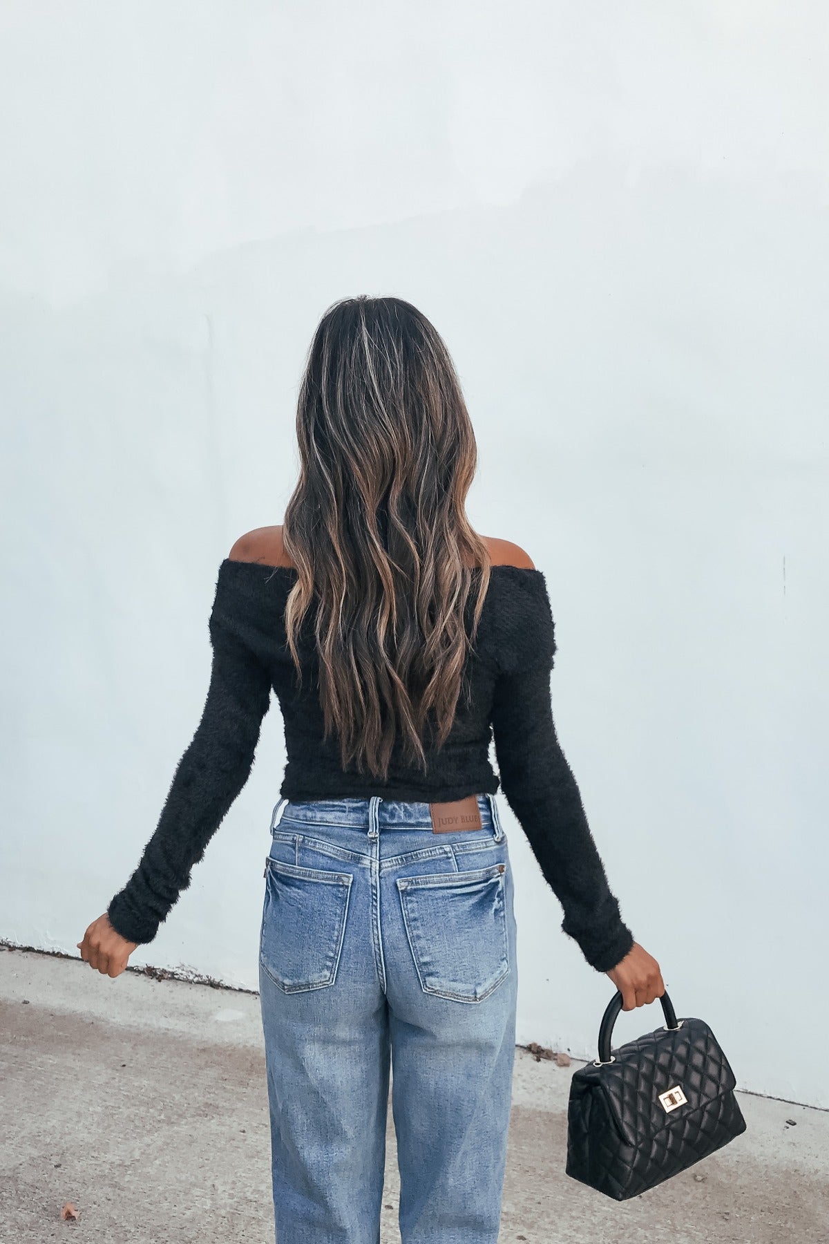 Woman with long wavy hair in a Black Fuzzy Knit Off-Shoulder Top and jeans, holding a black purse, stands facing away.
