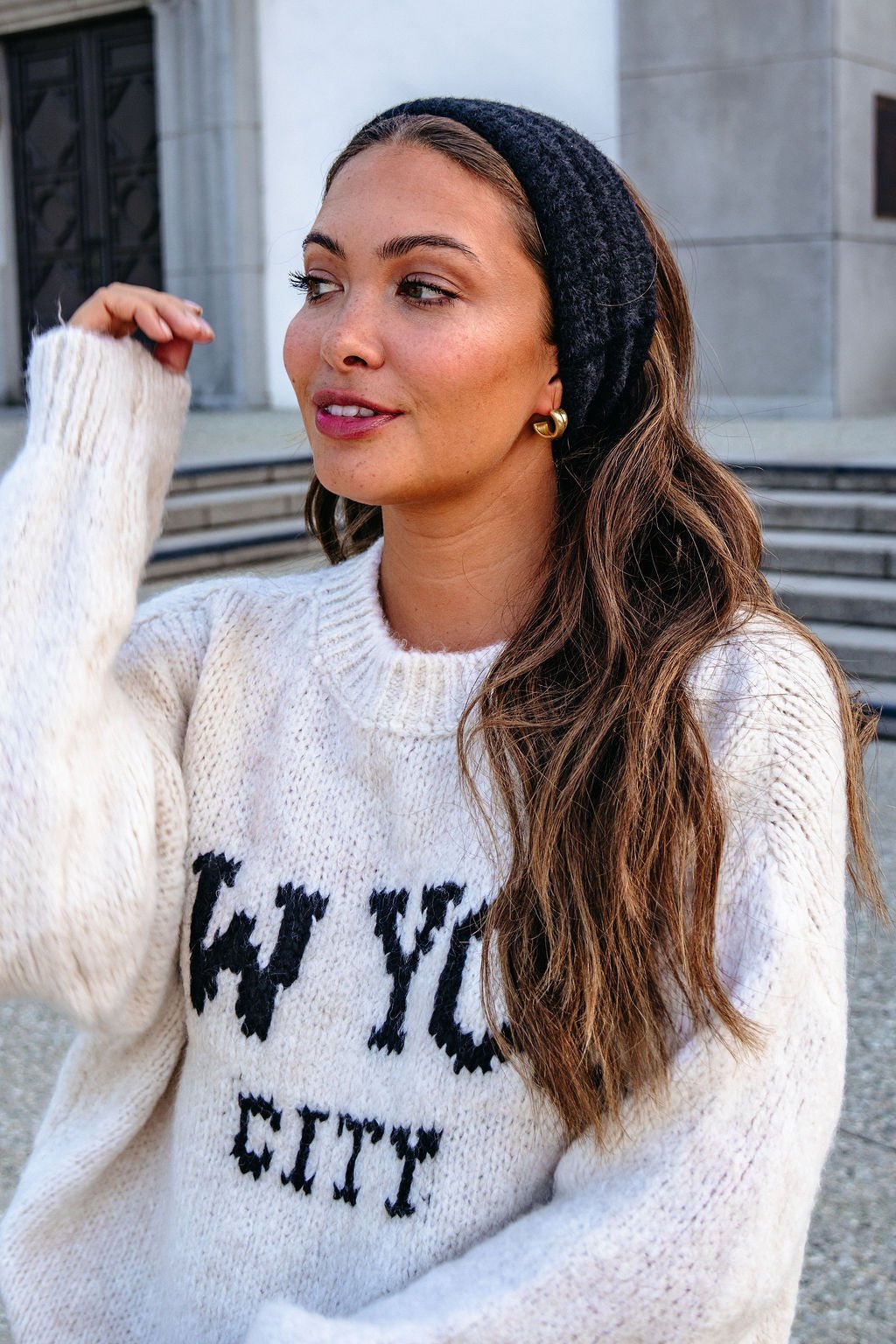 Woman with long brown hair wears a Black Fuzzy Soft Cable Knit Headband and a white NYC sweater, standing outdoors and smiling.