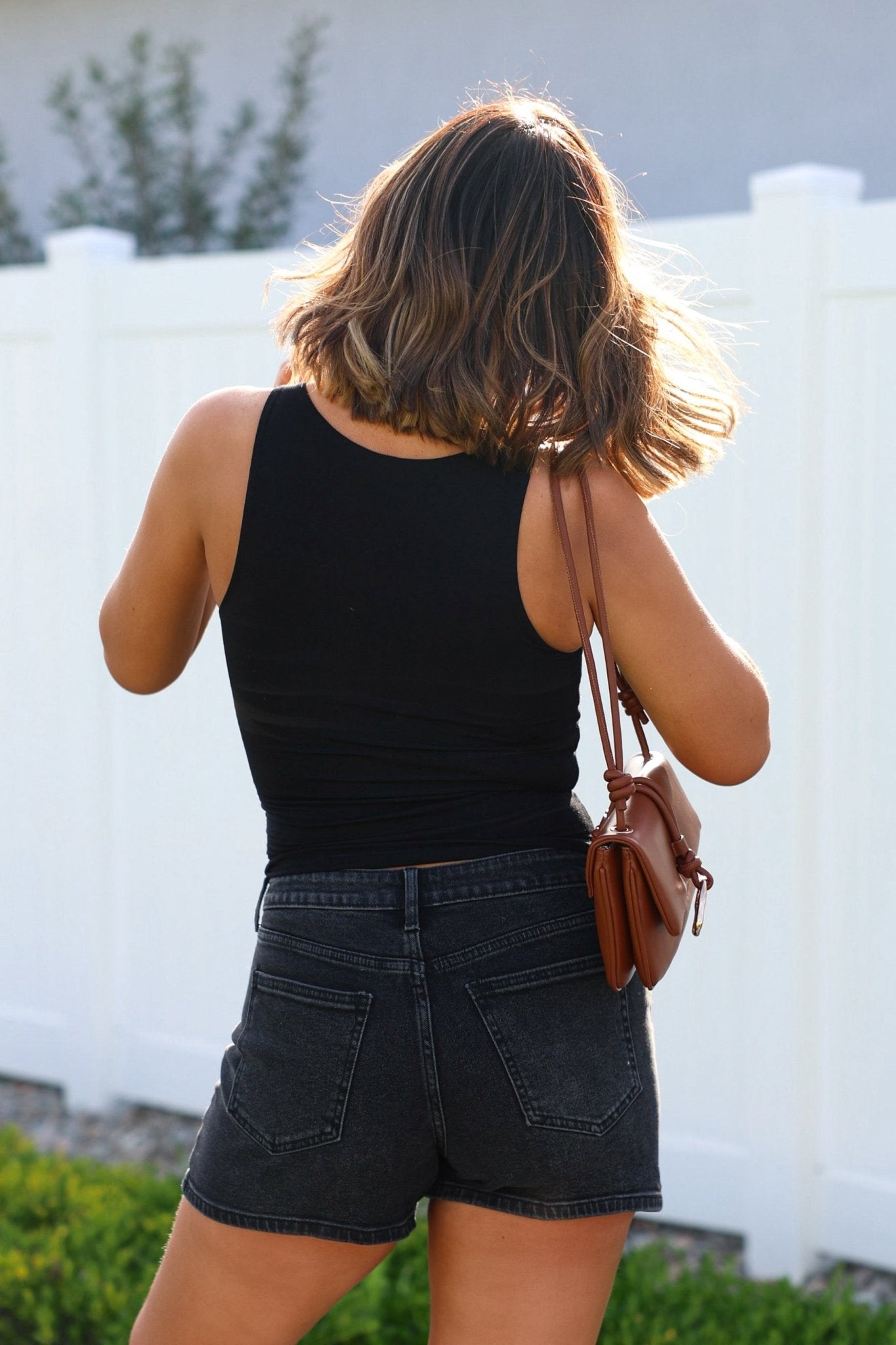A woman in Black High Rise Classic Hem Shorts and a black tank top stands outside by a white fence, carrying a brown purse.